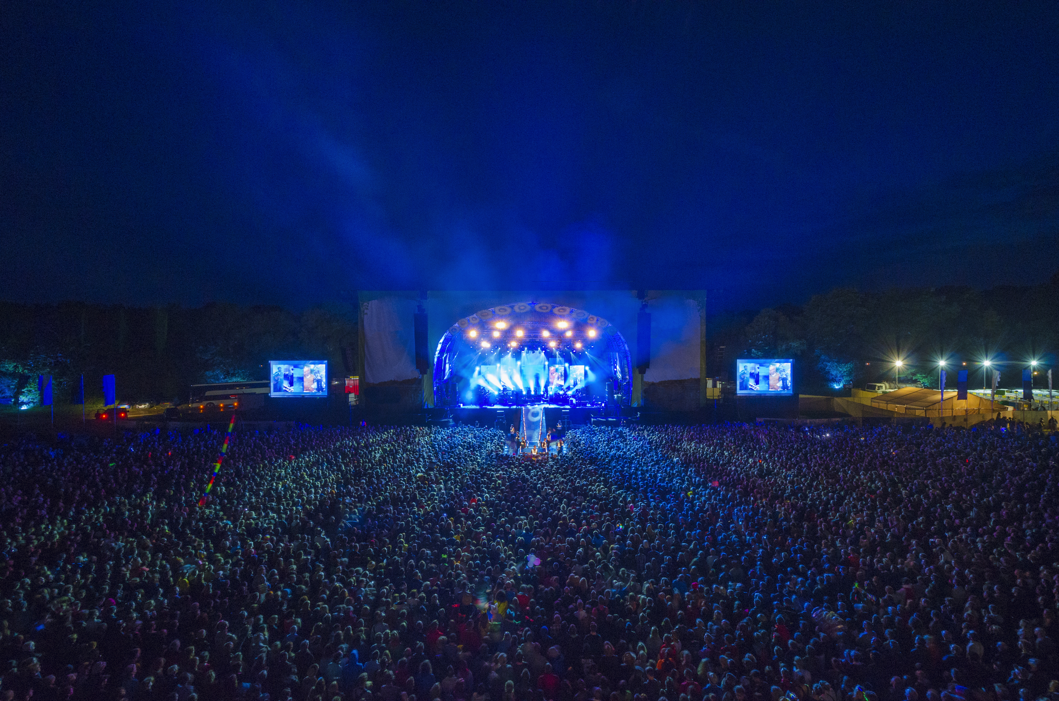 Large open-air concert with a packed audience, stage lights illuminating performers