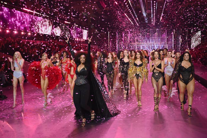 A group of models in various lingerie outfits walk a runway adorned with confetti and bright lights at a fashion show