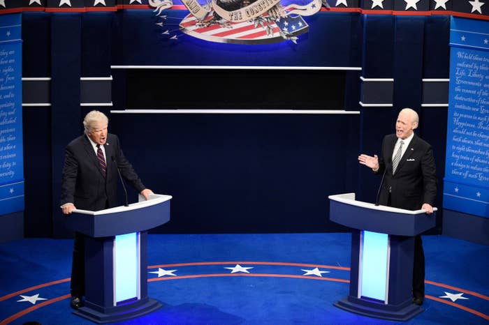 Two men, Alec Baldwin as Trump, and Jim Carrey as Biden, stand at podiums during a debate scene on a stage with patriotic decor