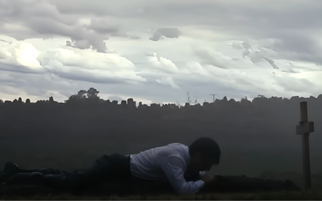 A person in a white shirt lies face down in a misty field near a wooden cross, under a cloudy sky, conveying a somber atmosphere