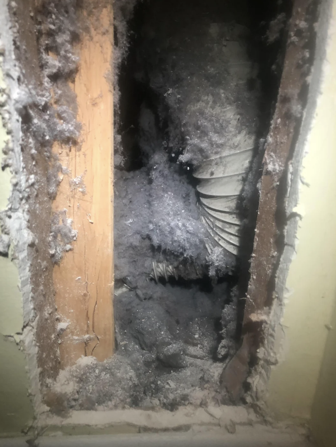 Dusty dryer vent clogged with lint inside a wall, showing the inner pipe and wooden framing