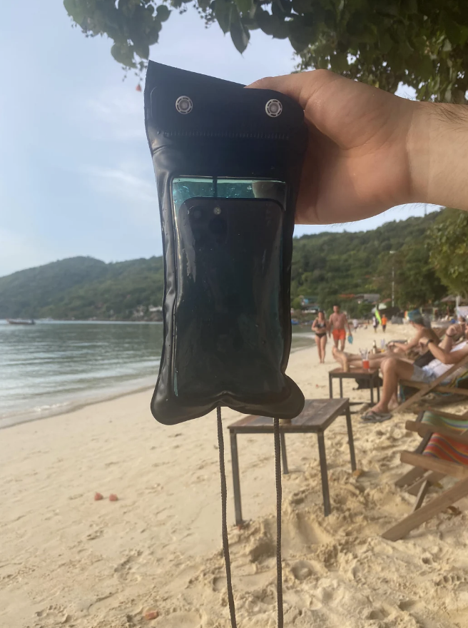 Hand holding a waterproof phone pouch on a beach, with people lounging and trees in the background