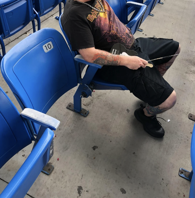 Person sitting in a stadium seat, wearing a band t-shirt, black shorts, and sneakers. Tattoos visible on arms and legs. Face not shown