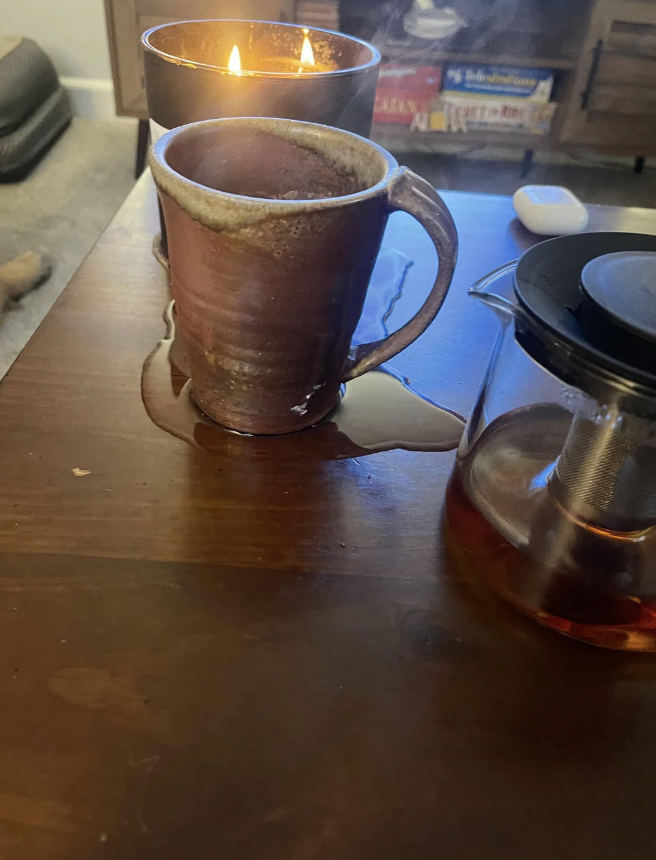 A ceramic mug on a wooden table with spilled liquid, next to a lit candle and a teapot. Board games in the background. Cozy setting