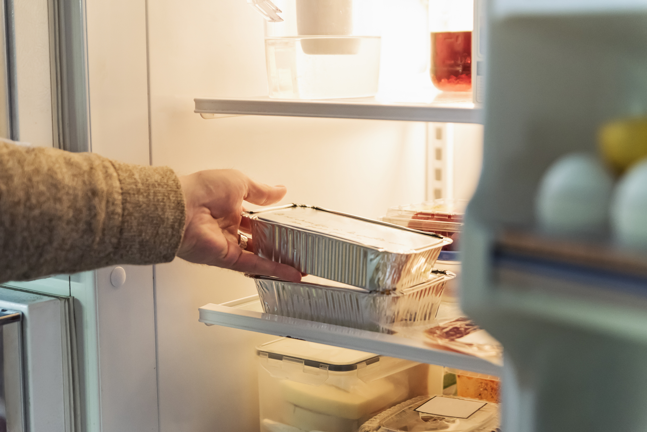 A hand places a foil container inside a refrigerator, surrounded by various food items and containers