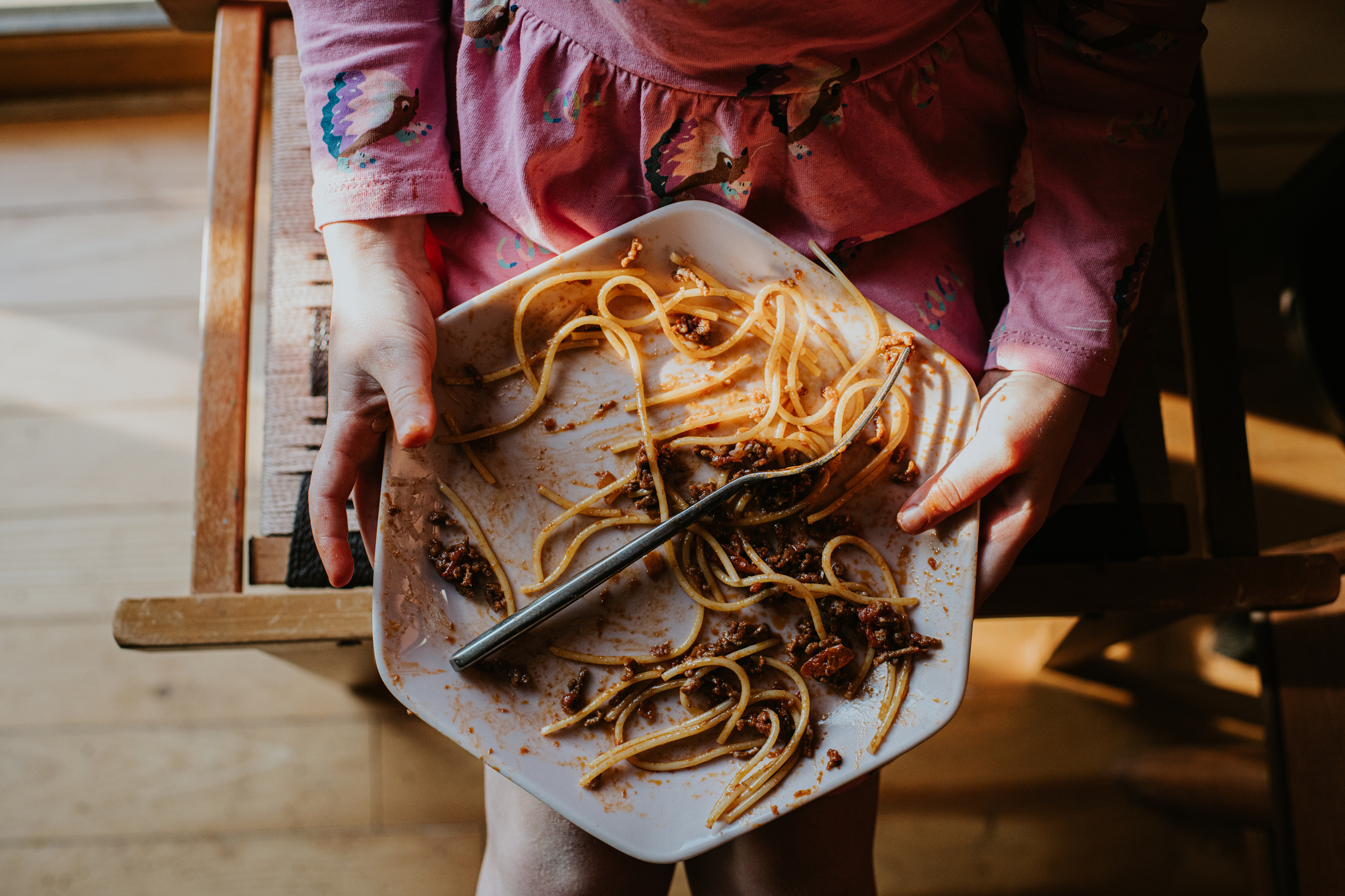 Child in a pink dress holding a plate with spaghetti and sauce in their lap