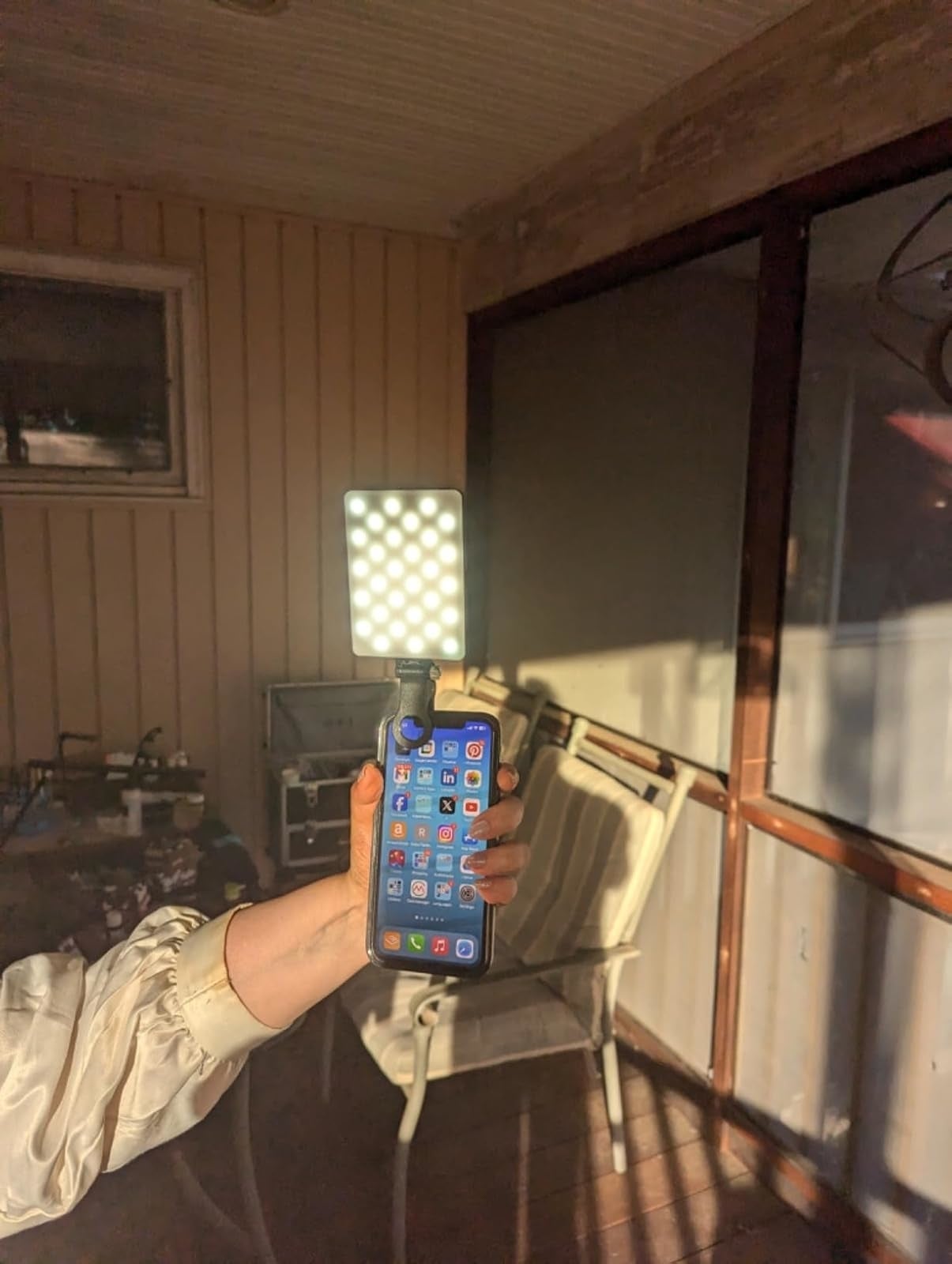 Person holding a smartphone with apps visible, attached to a square LED light. Background shows a porch with a chair and table