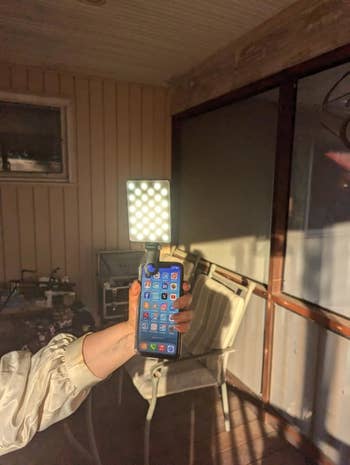 Person holding a smartphone with apps visible, attached to a square LED light. Background shows a porch with a chair and table