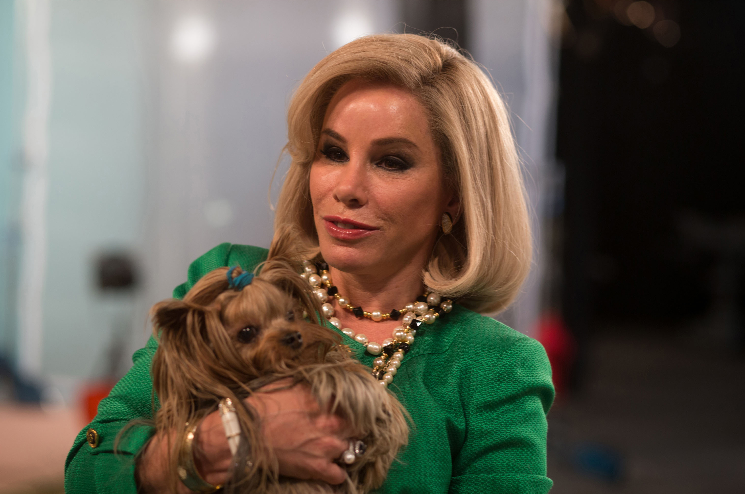 Melissa in a bob wig,  shirt with shoulder pads, and beaded necklace, holding a Yorkie