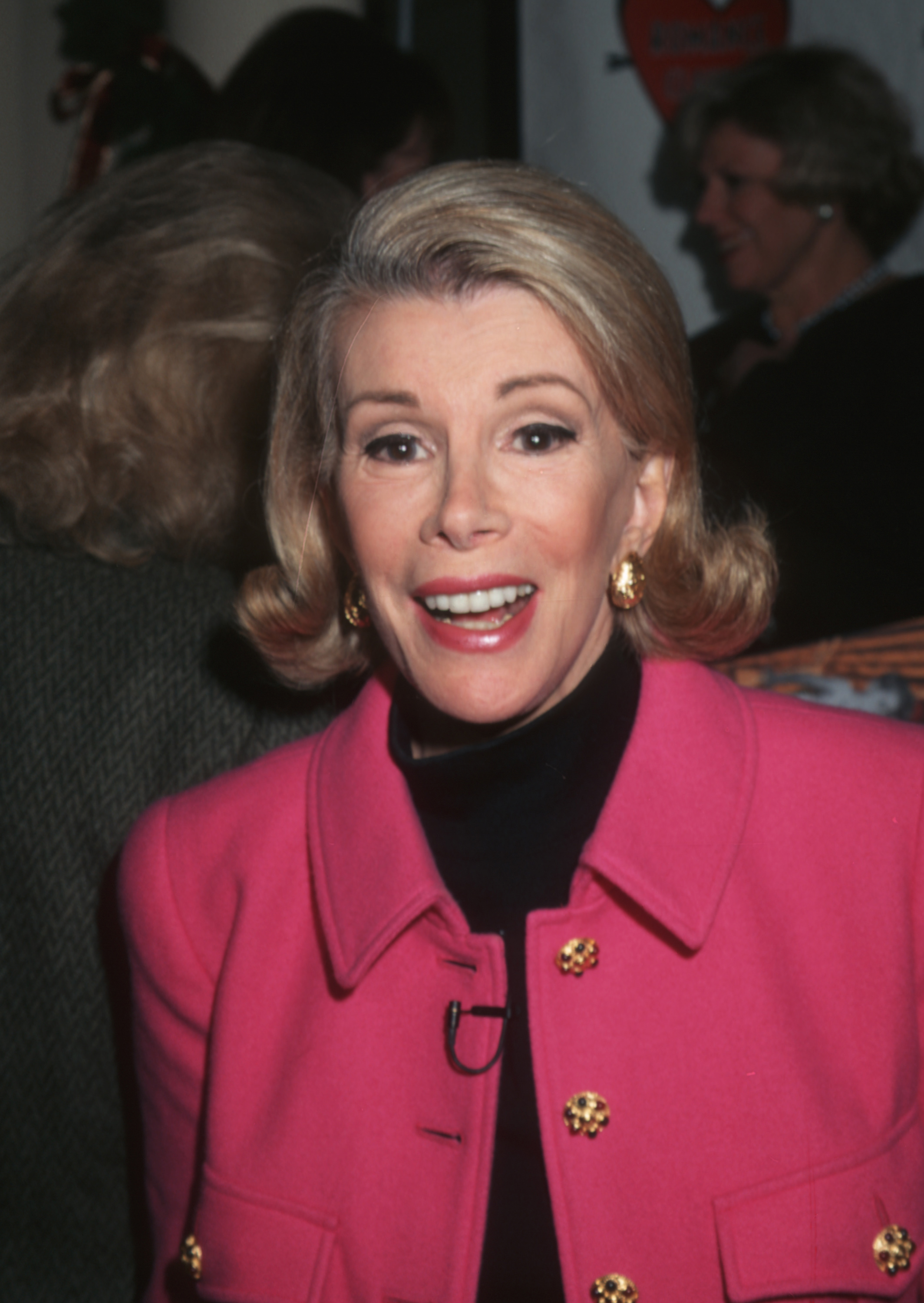 Joan Rivers with a curled bob haircut, wearing a brightly colored coat with metallic buttons and matching earrings