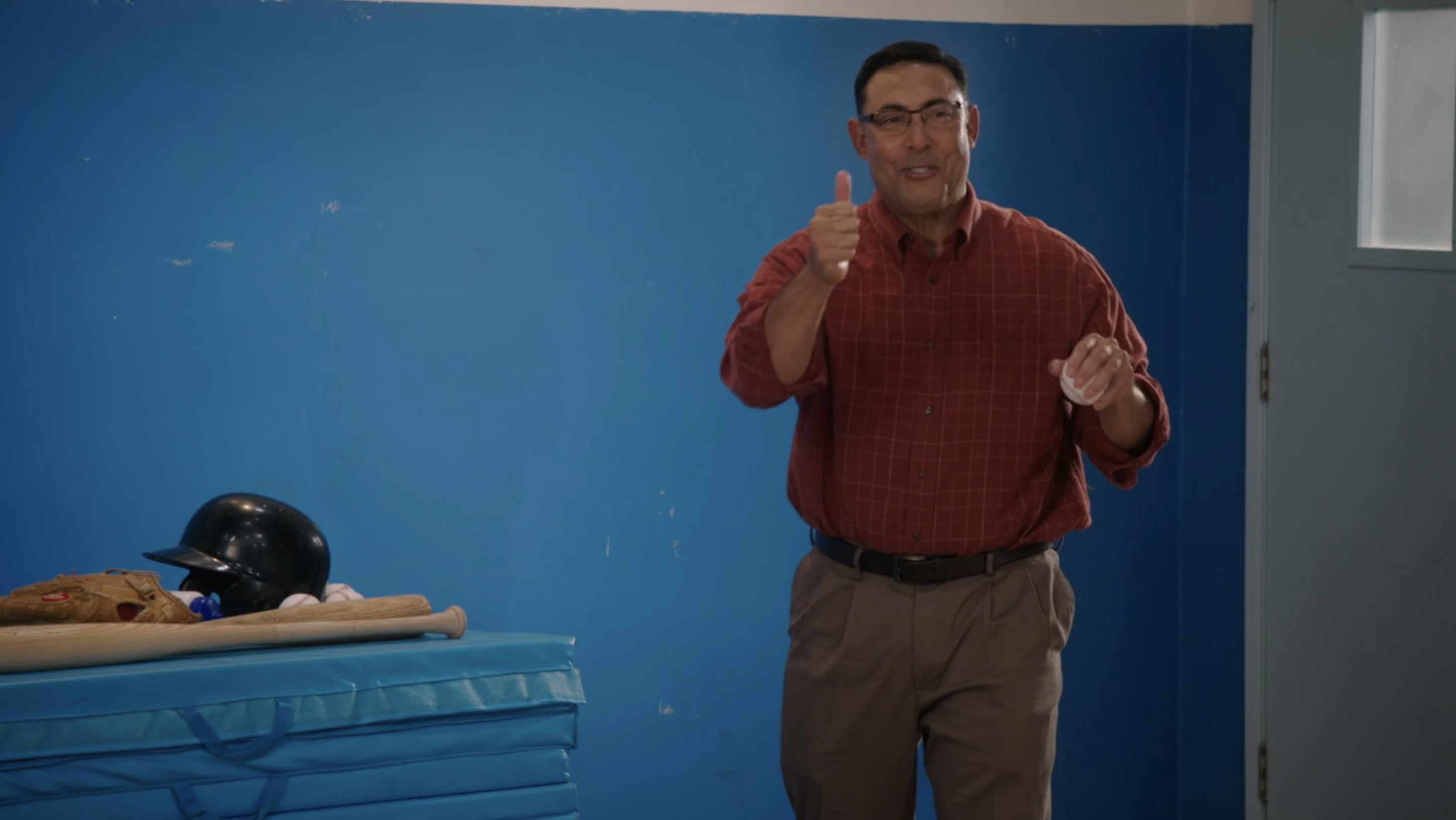 Rubén Jr in a casual plaid shirt gives a thumbs-up in a room with sports equipment