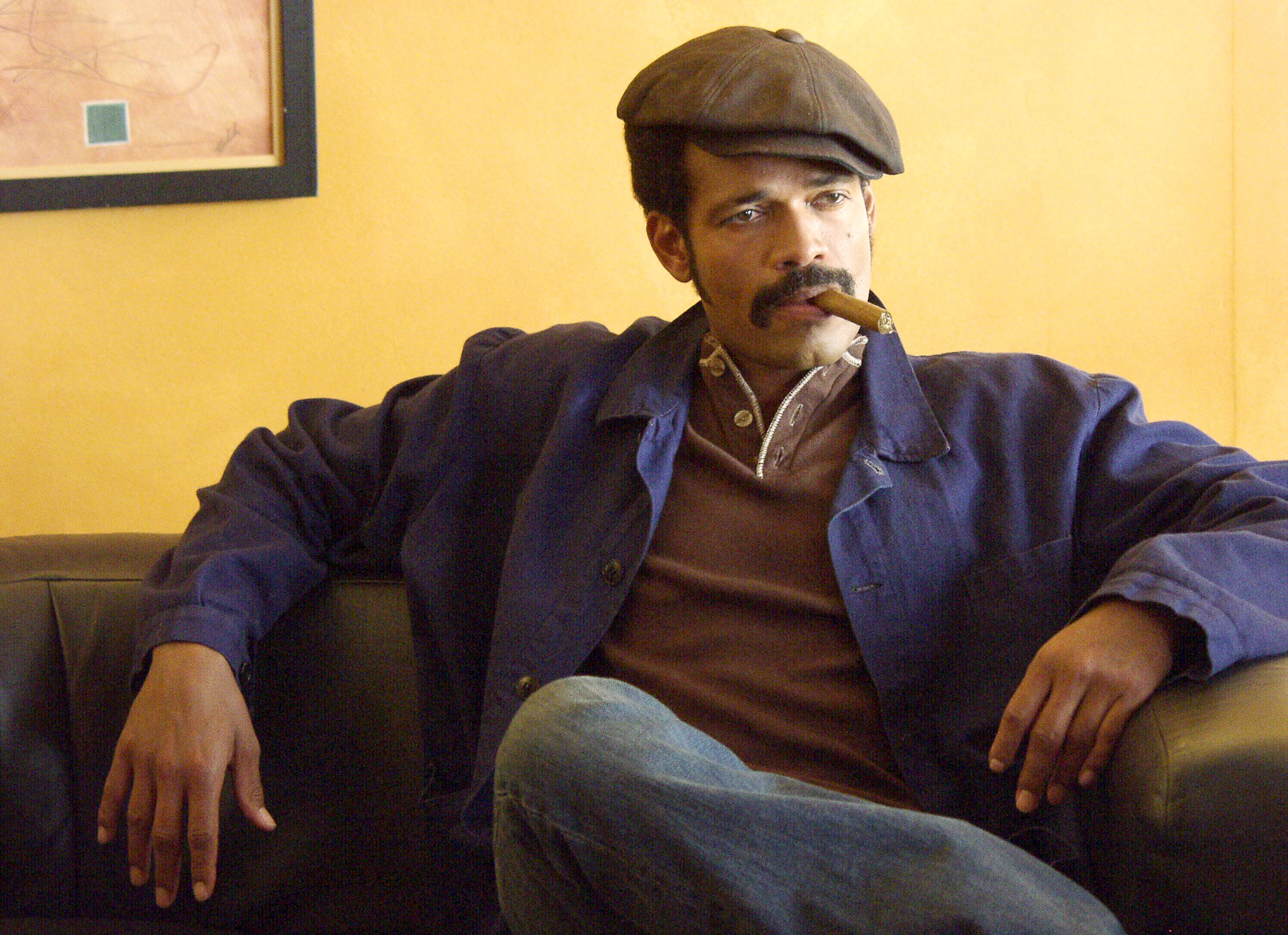 Mario wearing a jacket over a polo and jeans, with a newsboy cap and cigar. He&#x27;s lounging on a couch with his legs crossed