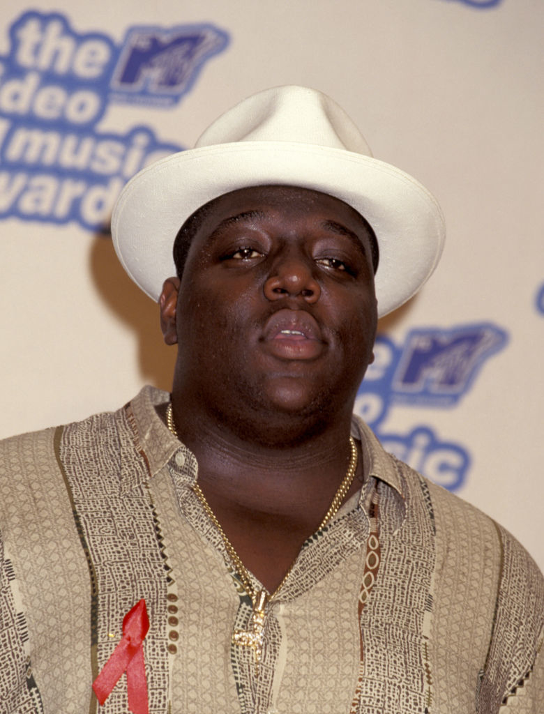 Biggie Smalls in a patterned shirt with a ribbon pinned to it, also wearing a gold chain and a hat