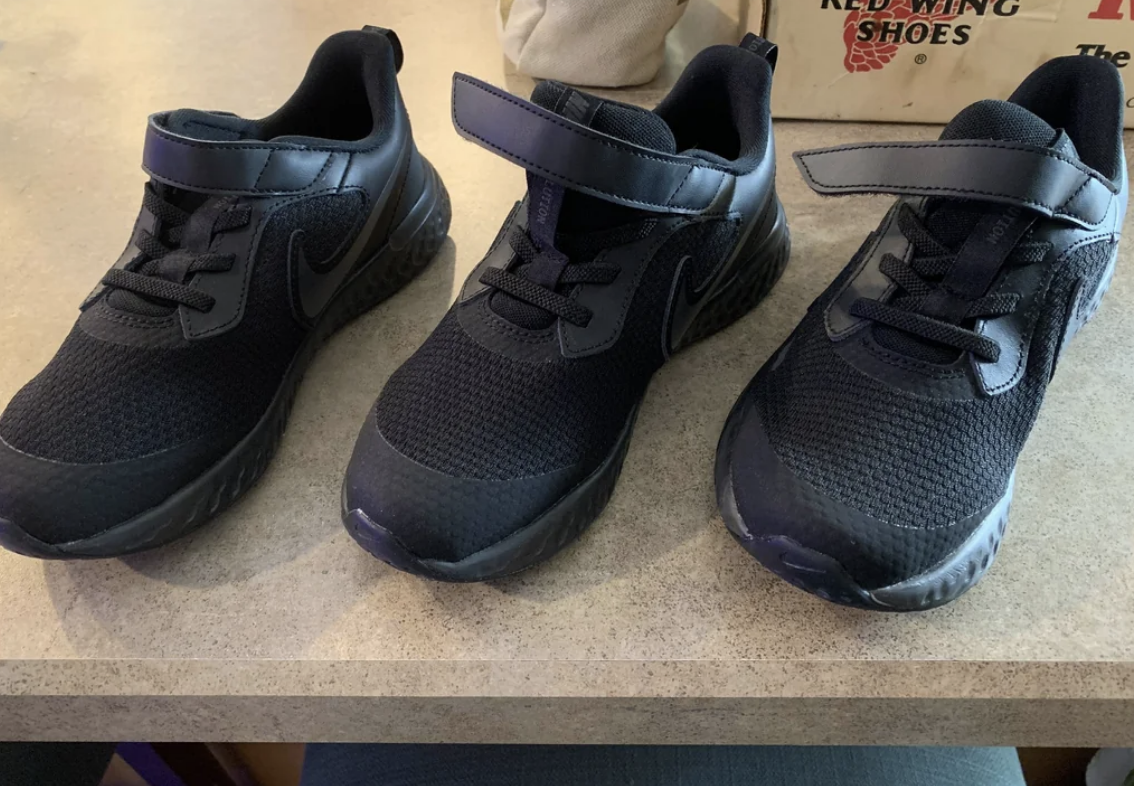 Three similar black athletic shoes on a countertop, two are left shoes and one is right. They are lace-ups with Velcro straps