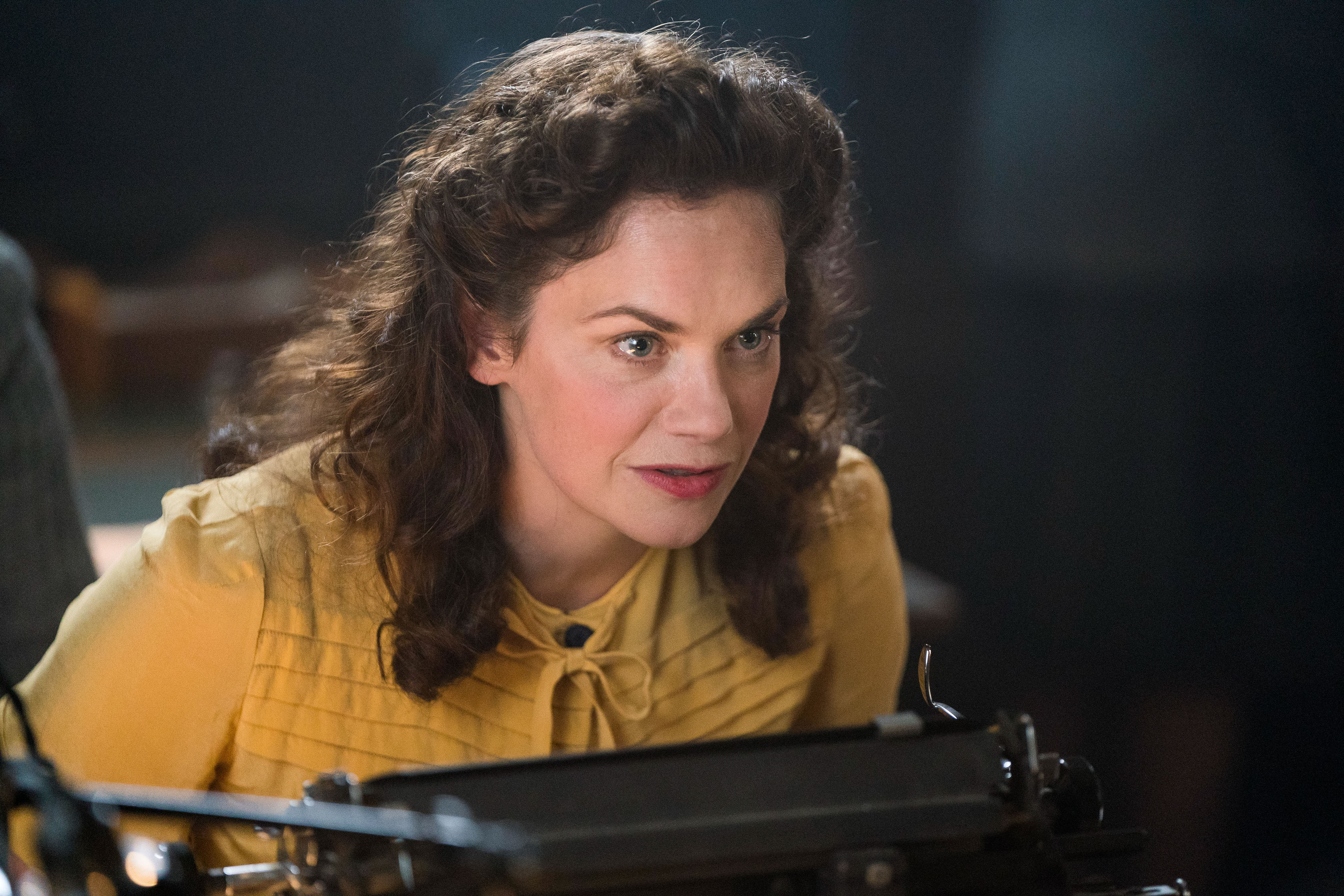 Alison with curly hair wears a vintage-style blouse, leaning intently toward a typewriter in a scene