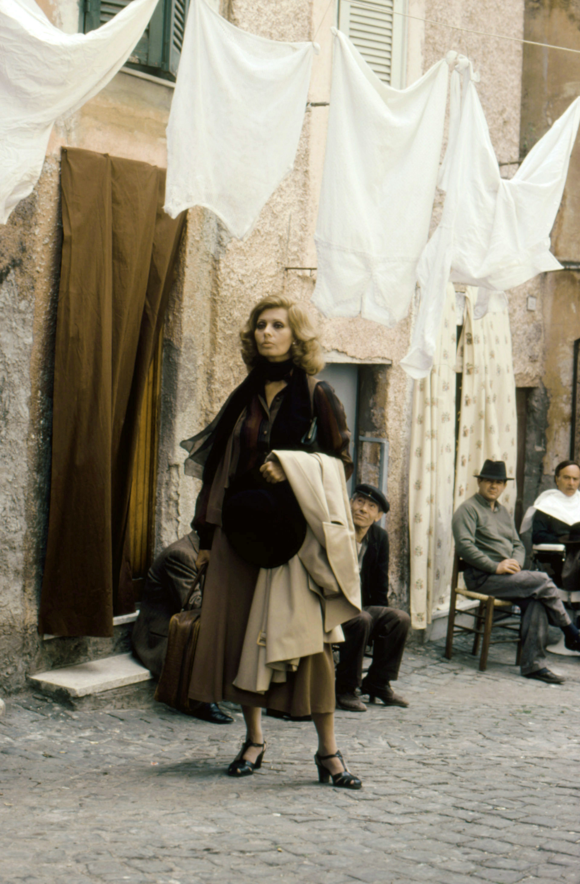 Sophia stands on a cobblestone street wearing a classic trench coat and shoulder bag, while people sit and watch in the background