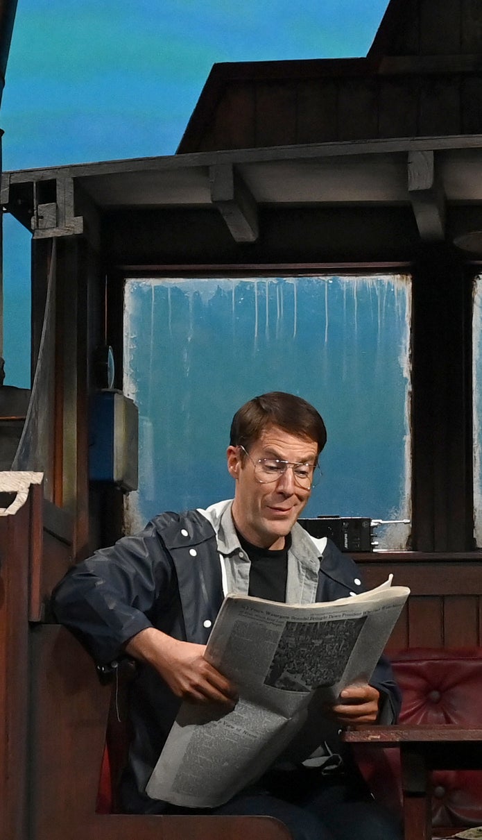 Ian on stage in a jacket and ballcap, sitting at a table