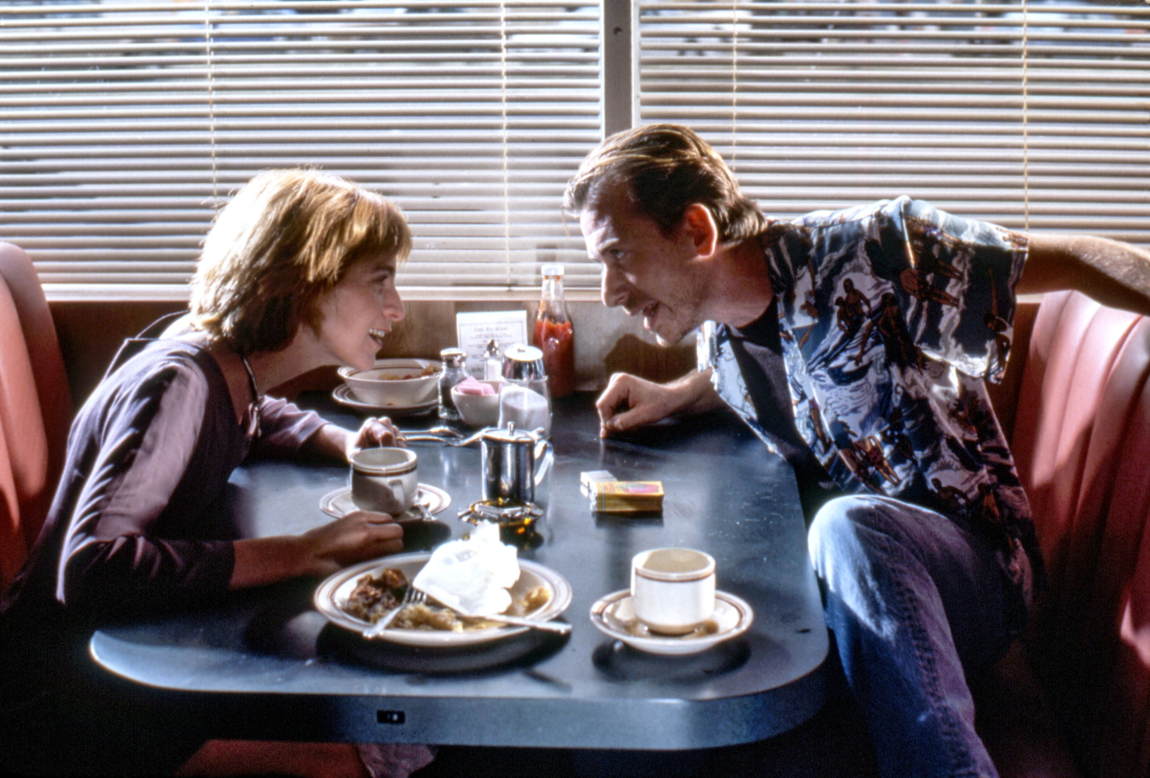 A man and a woman sit across from each other in a diner booth, leaning in for a conversation over coffee