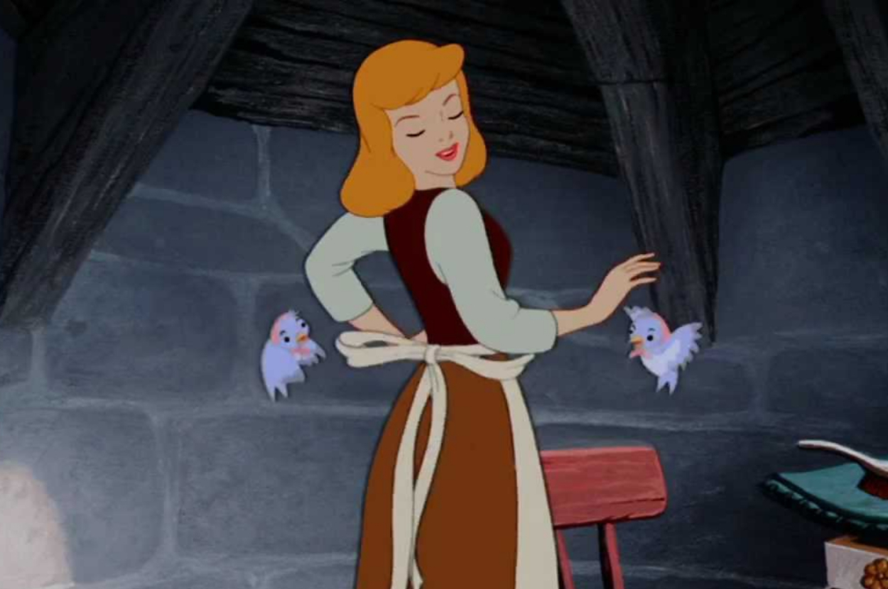 Cinderella in a brown apron smiles with birds fluttering around her inside a quaint attic