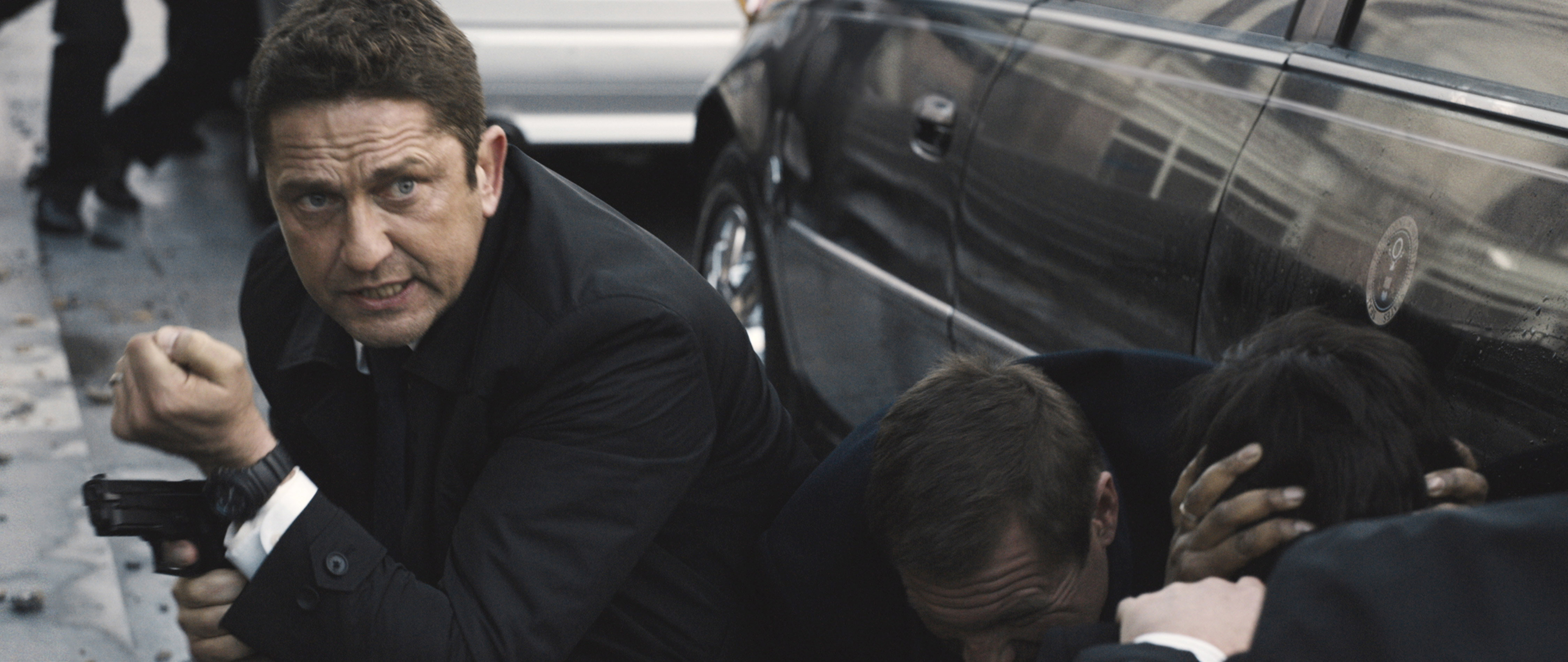 Man in a suit holding a gun crouches next to two others hiding, by a car, in an action scene