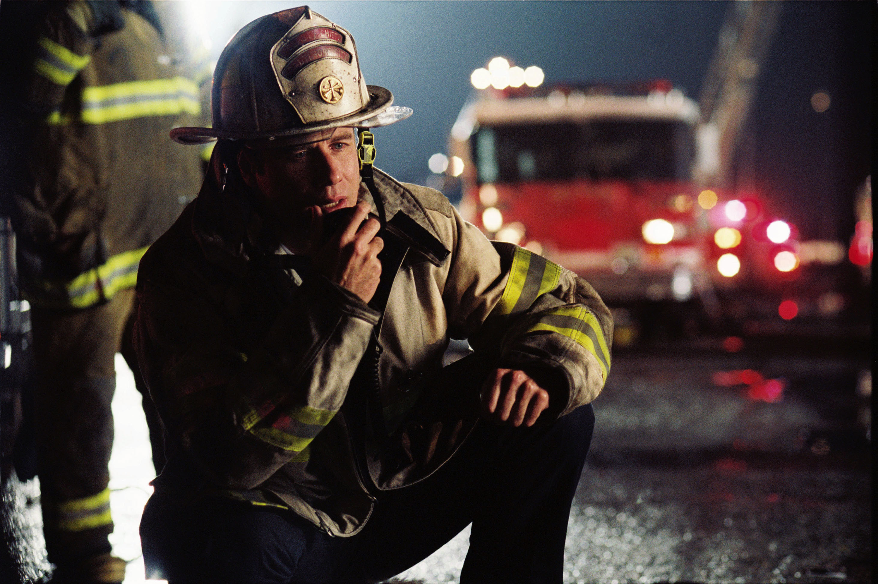 Firefighter in gear communicates via radio at nighttime scene with blurred emergency vehicle lights in background