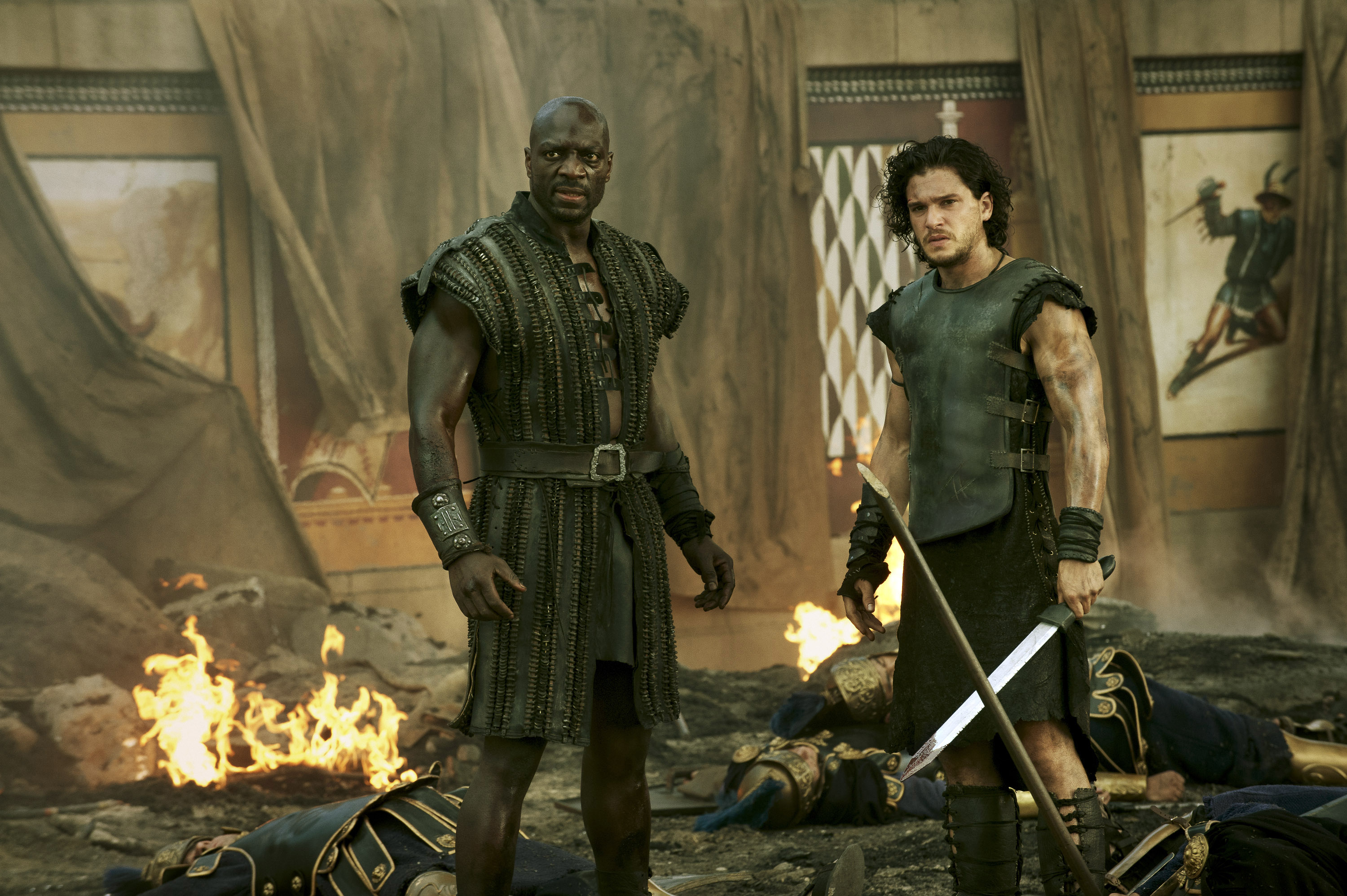 Two men in warrior costumes stand amidst ruins and flames, holding swords