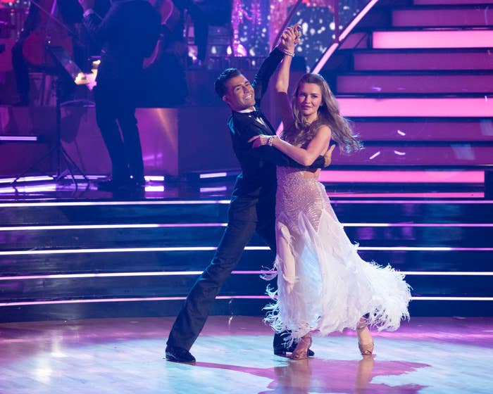 Ezra Sosa and Anna Delvey in elegant dance attire perform a ballroom routine on a stage with steps and lighting in the background