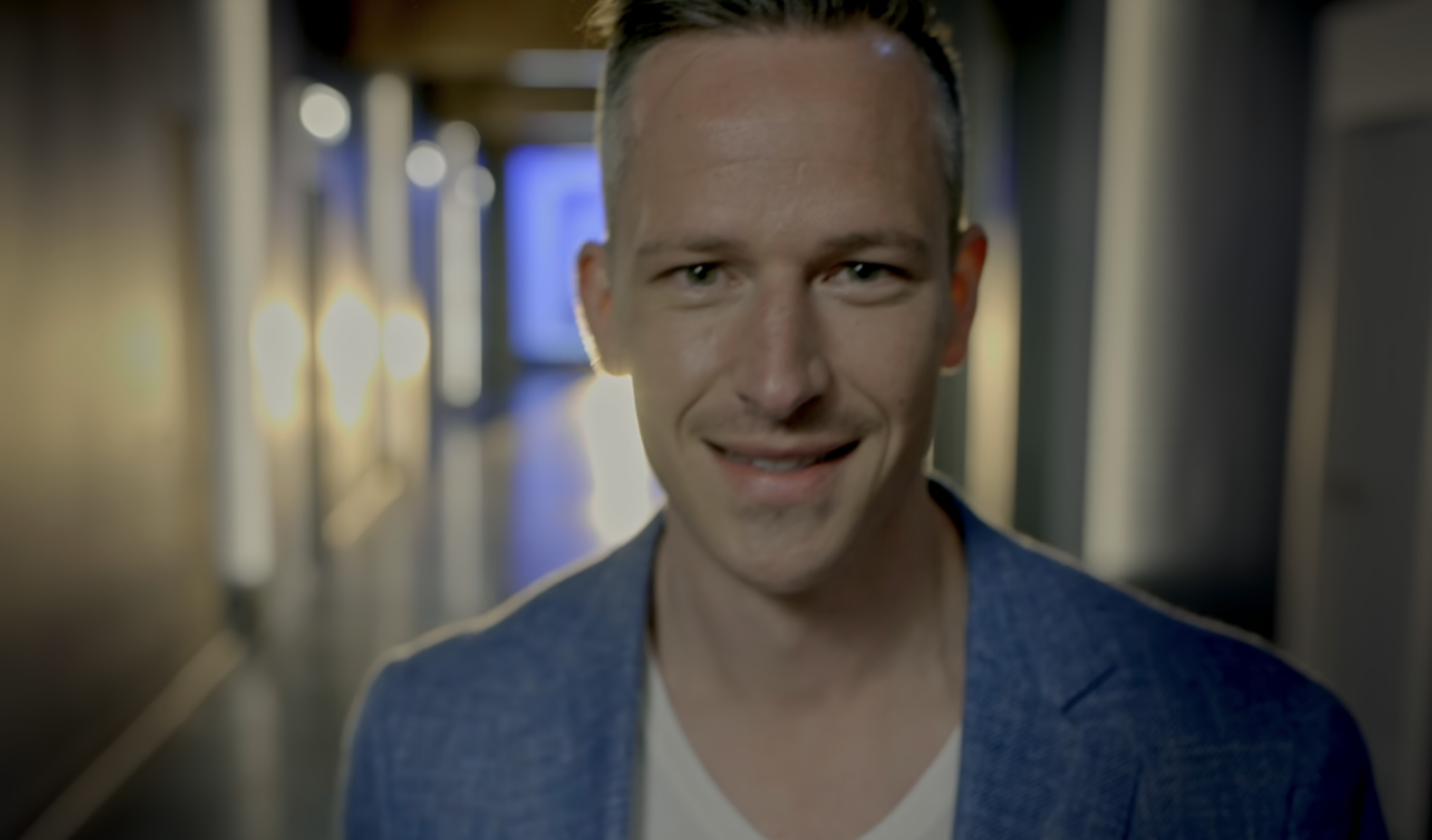 Jeremy, in a blazer and t-shirt, smiles while walking down a hallway filled with doors