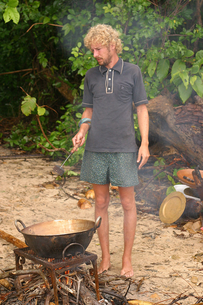 Jonny on beach in casual outfit cooking with large pot over fire