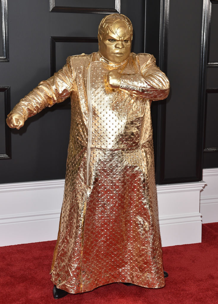 A person on a red carpet dressed in a full-length, gold metallic outfit with a textured design, appearing to strike a pose