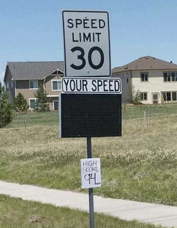 Roadside speed limit sign reads &quot;Speed Limit 30, Your Speed.&quot; Below, a handwritten sign says &quot;High Score 94.&quot;