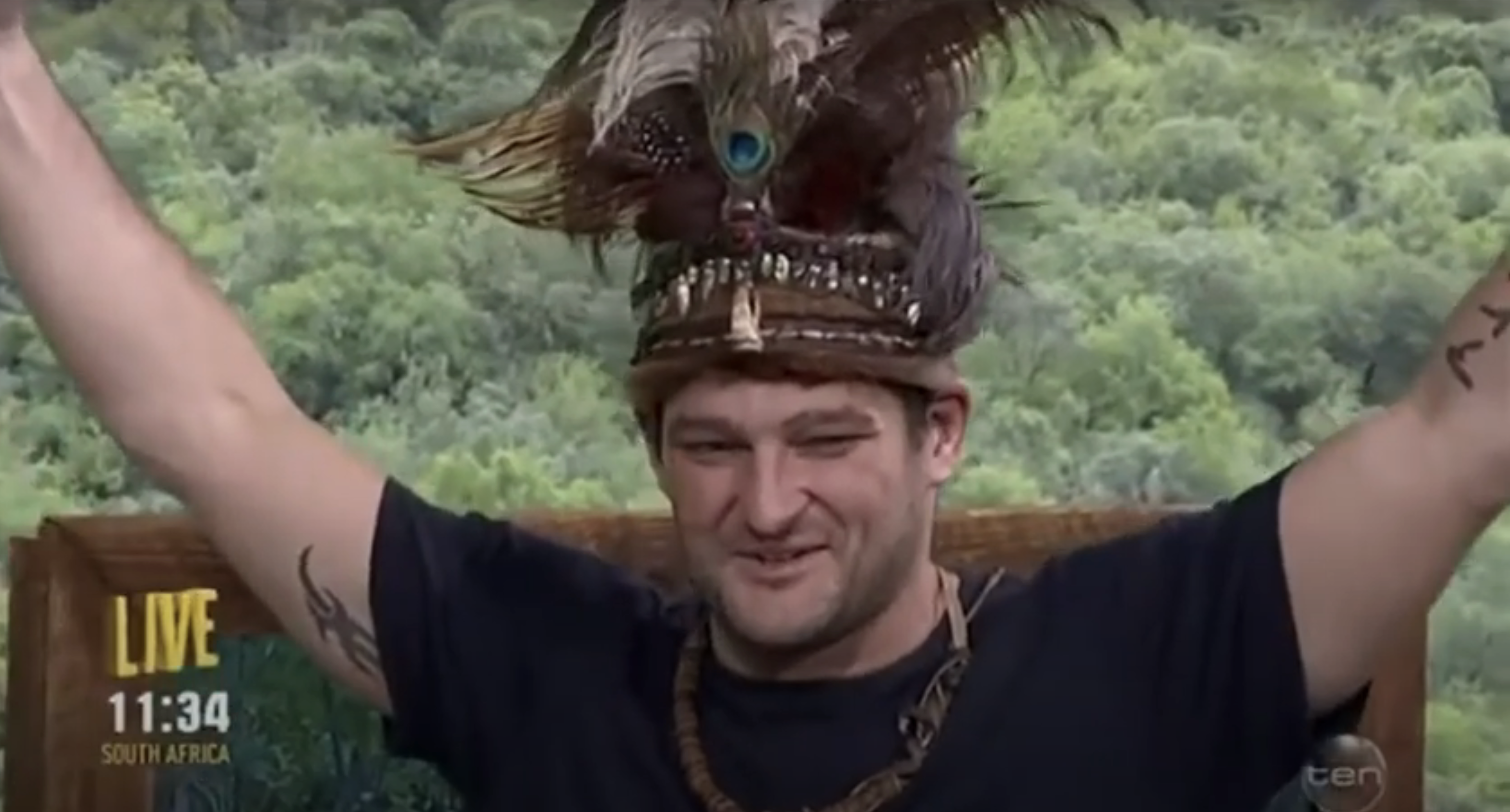 Fev on a live TV show wears a tribal-style headdress and raises his arms in triumph. Trees are visible in the background