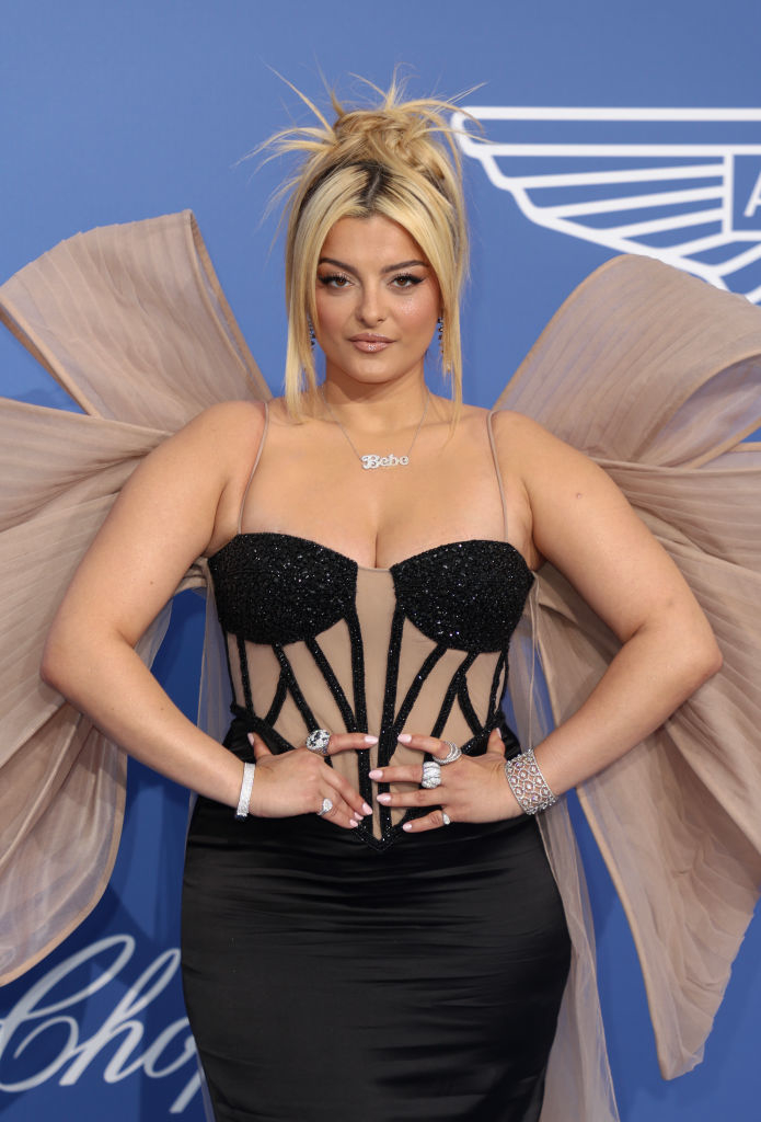 I don’t know who the person is, but they are on a red carpet wearing a black corset dress with sheer details and large bow accents