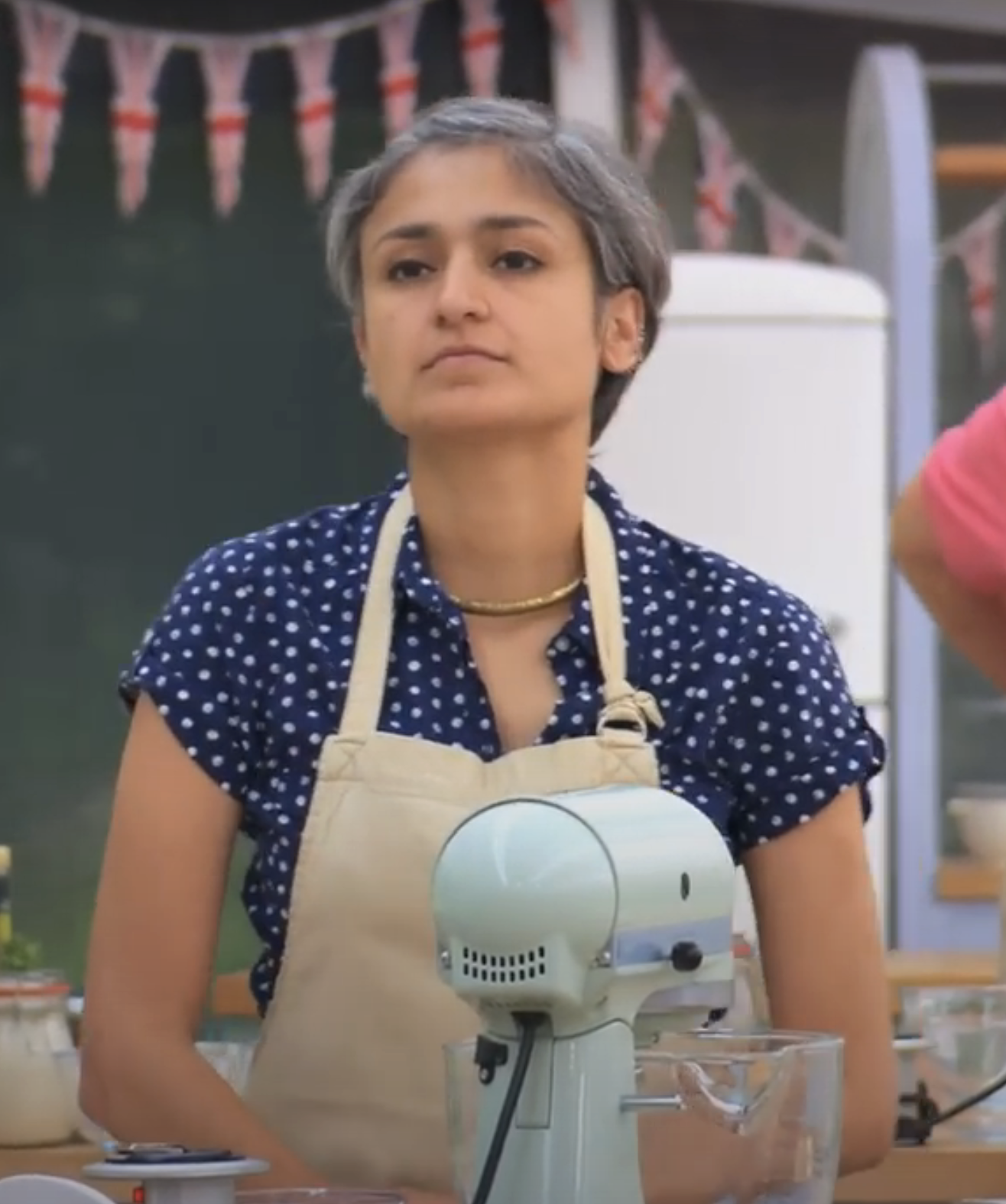 Chetna in a polka dot shirt and apron is standing in a kitchen with a mixer in front of her