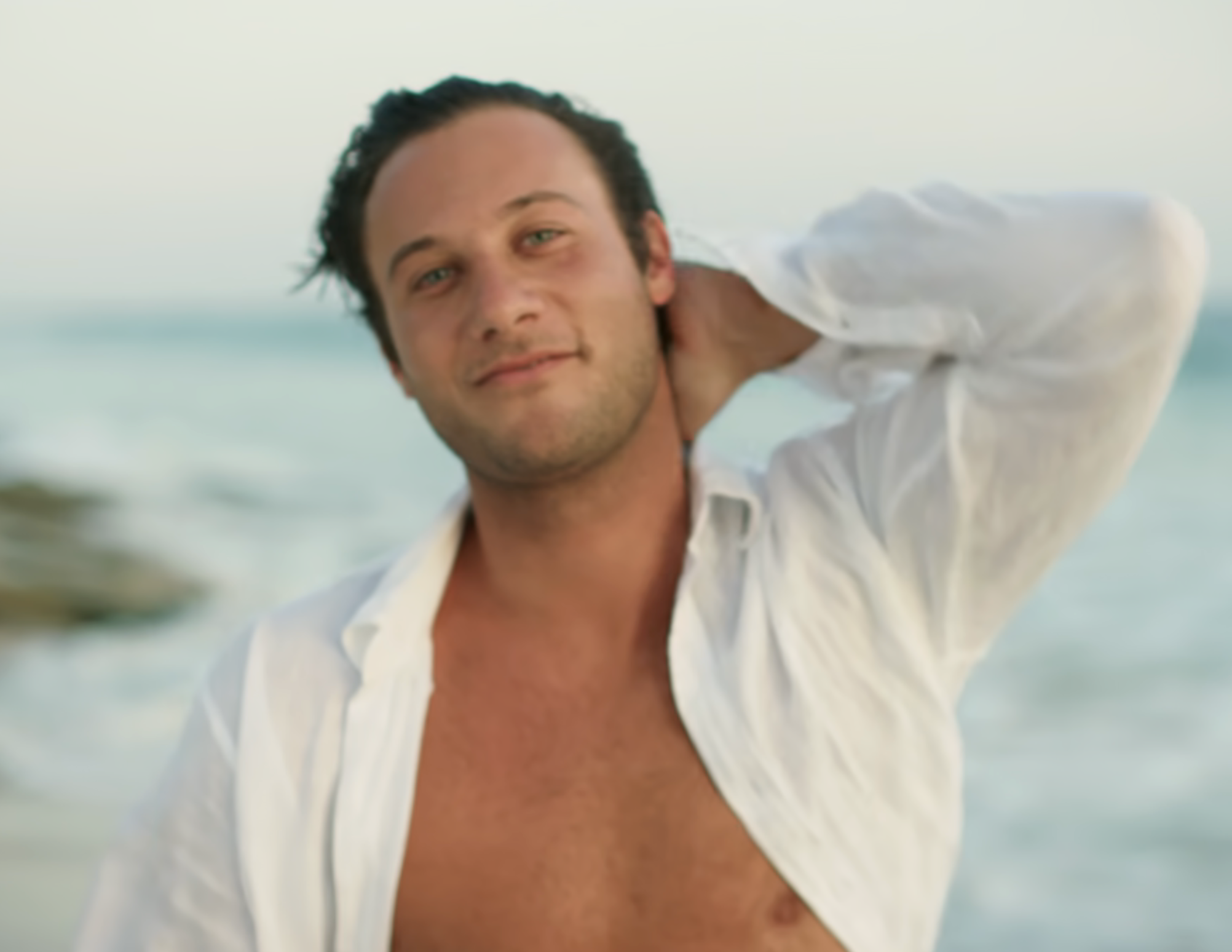 Bryce on a beach wearing an open shirt, smiling with a relaxed pose