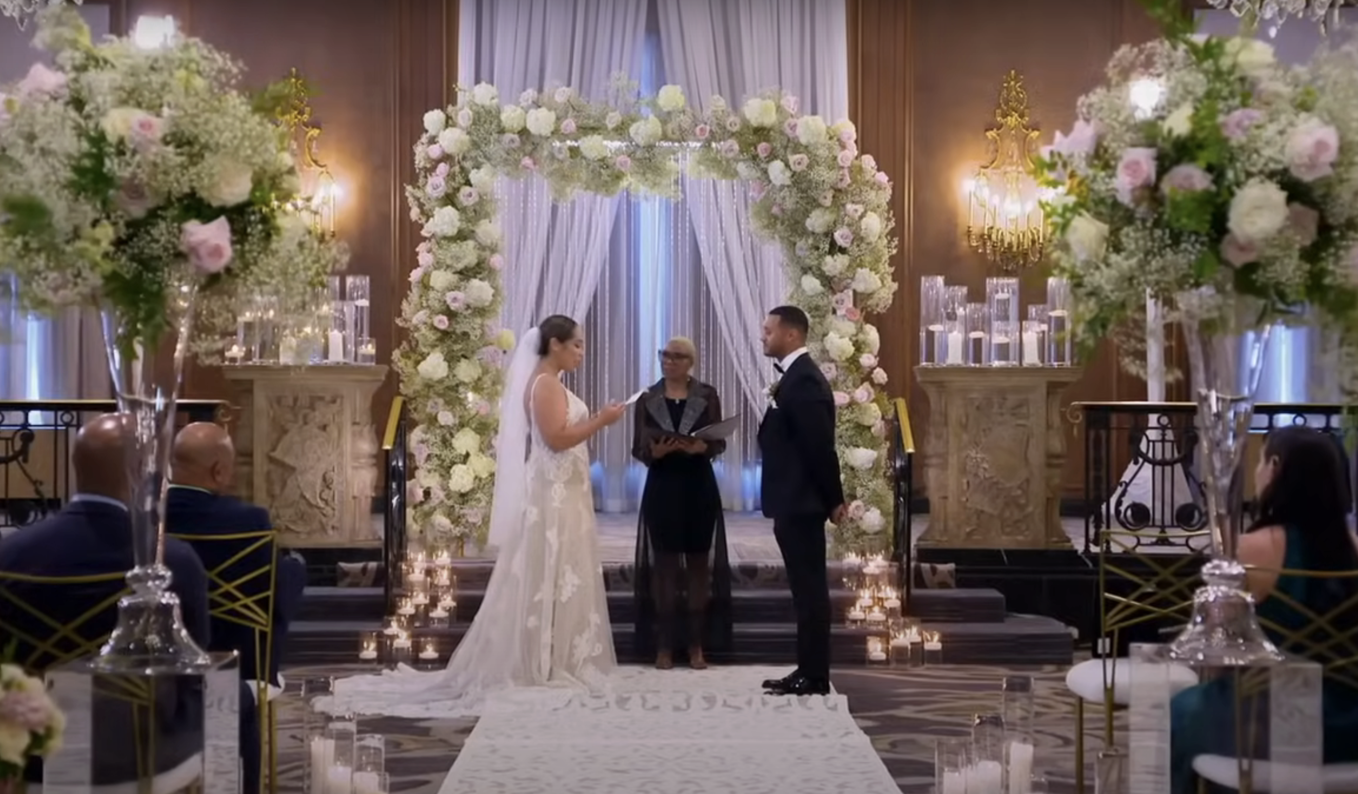 Camille in an elegant lace gown and Thomas in a tuxedo stand at the altar during a lavish indoor wedding ceremony