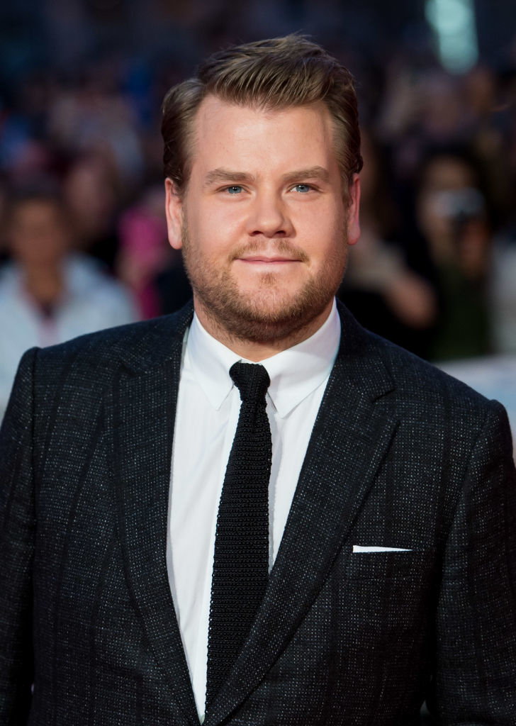 A person in a tailored suit and tie stands on a red carpet, smiling at the camera