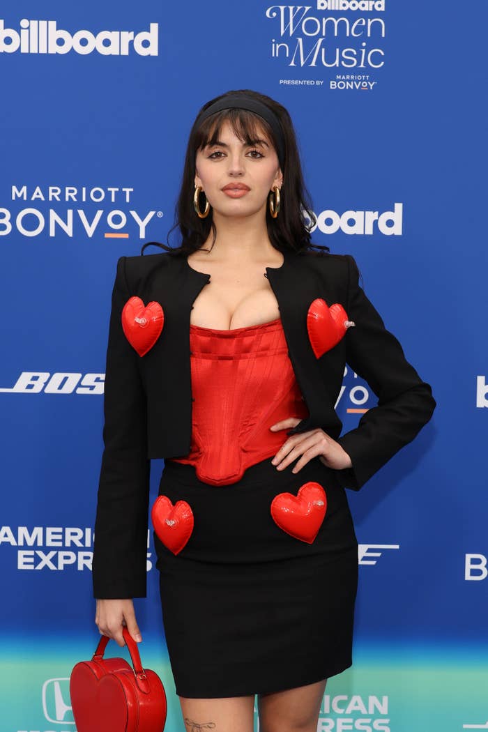 The person is on a red carpet, wearing a black jacket and skirt with red heart accents, and holding a heart-shaped purse