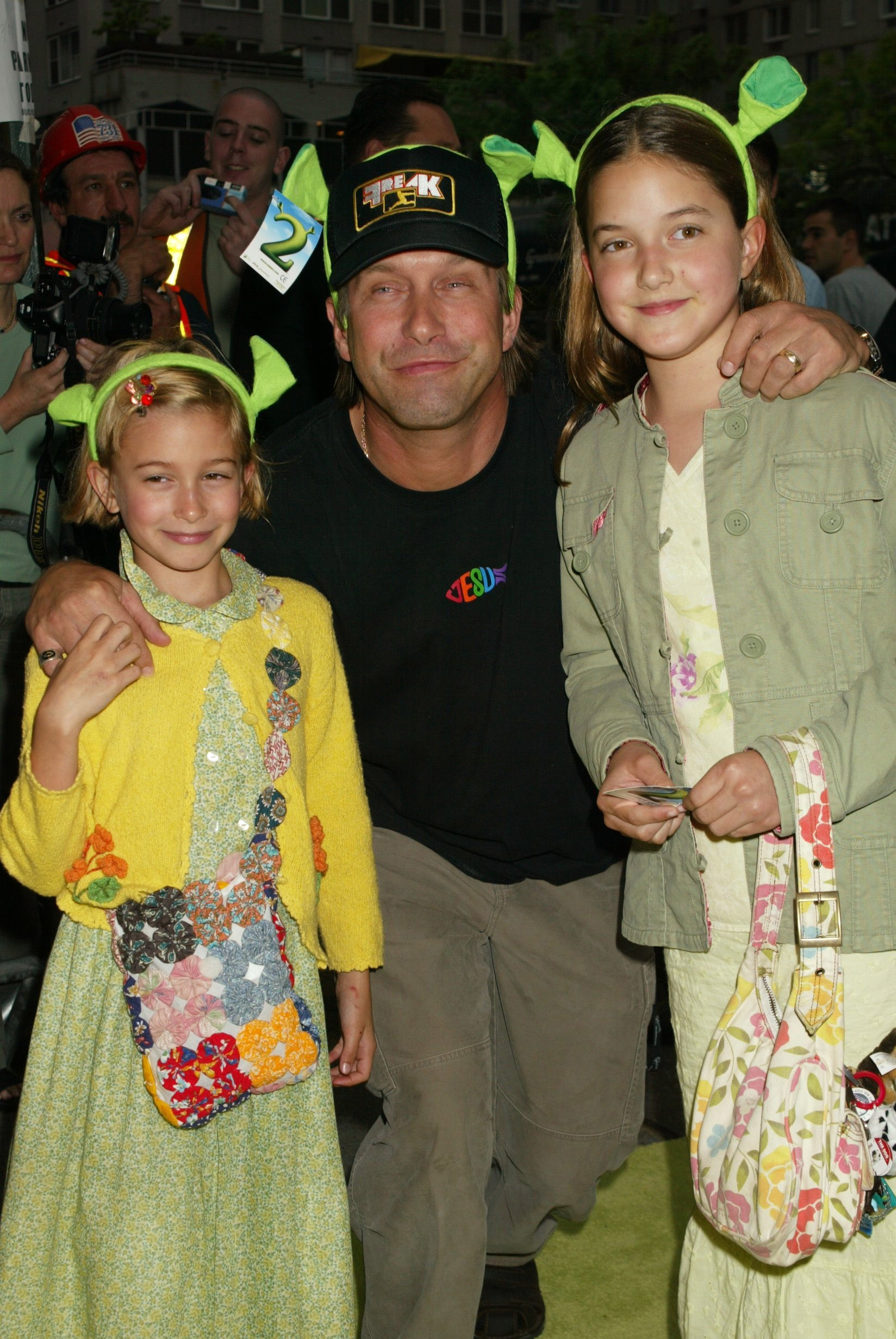 A man posing with two children at a public event. Children wear outfits with unique patterns. The man wears a cap and casual clothing. Celebrities unknown