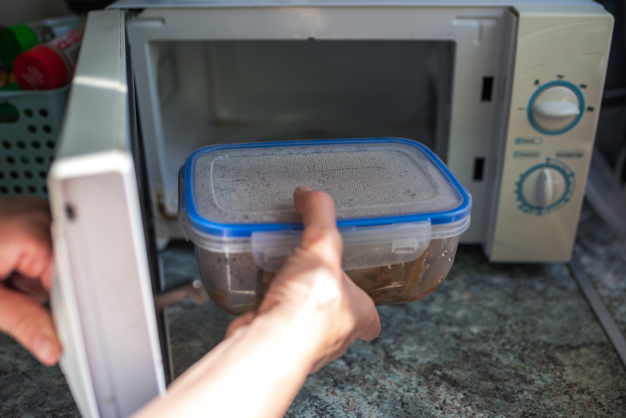 Person places a plastic container with food into an open microwave on a kitchen counter