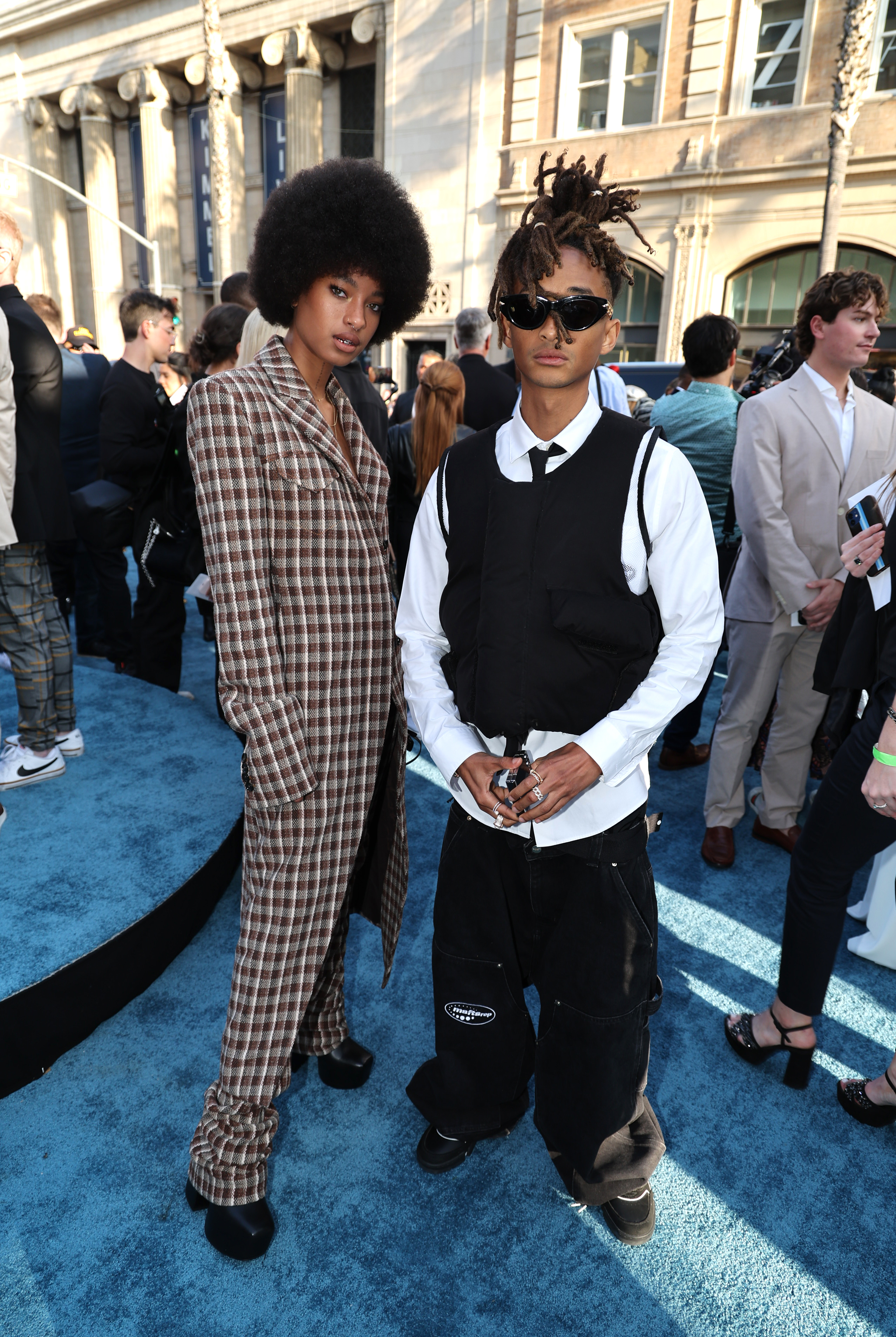 I don’t know who these people are, but they are posing on a blue carpet. One is wearing a plaid suit and the other a black vest and pants with sunglasses