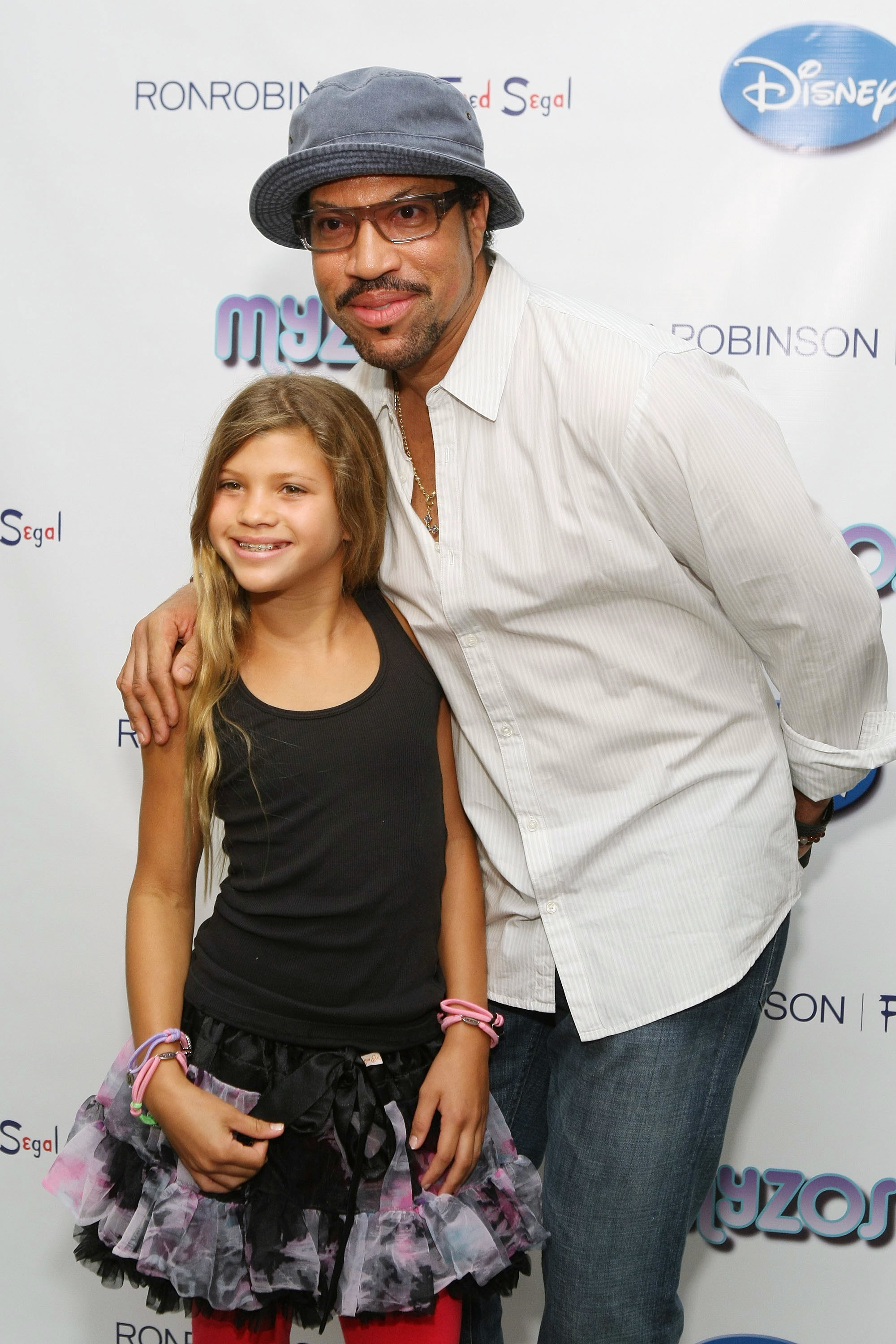 A man in a hat and glasses with a young girl on a red carpet event; both smiling at the camera