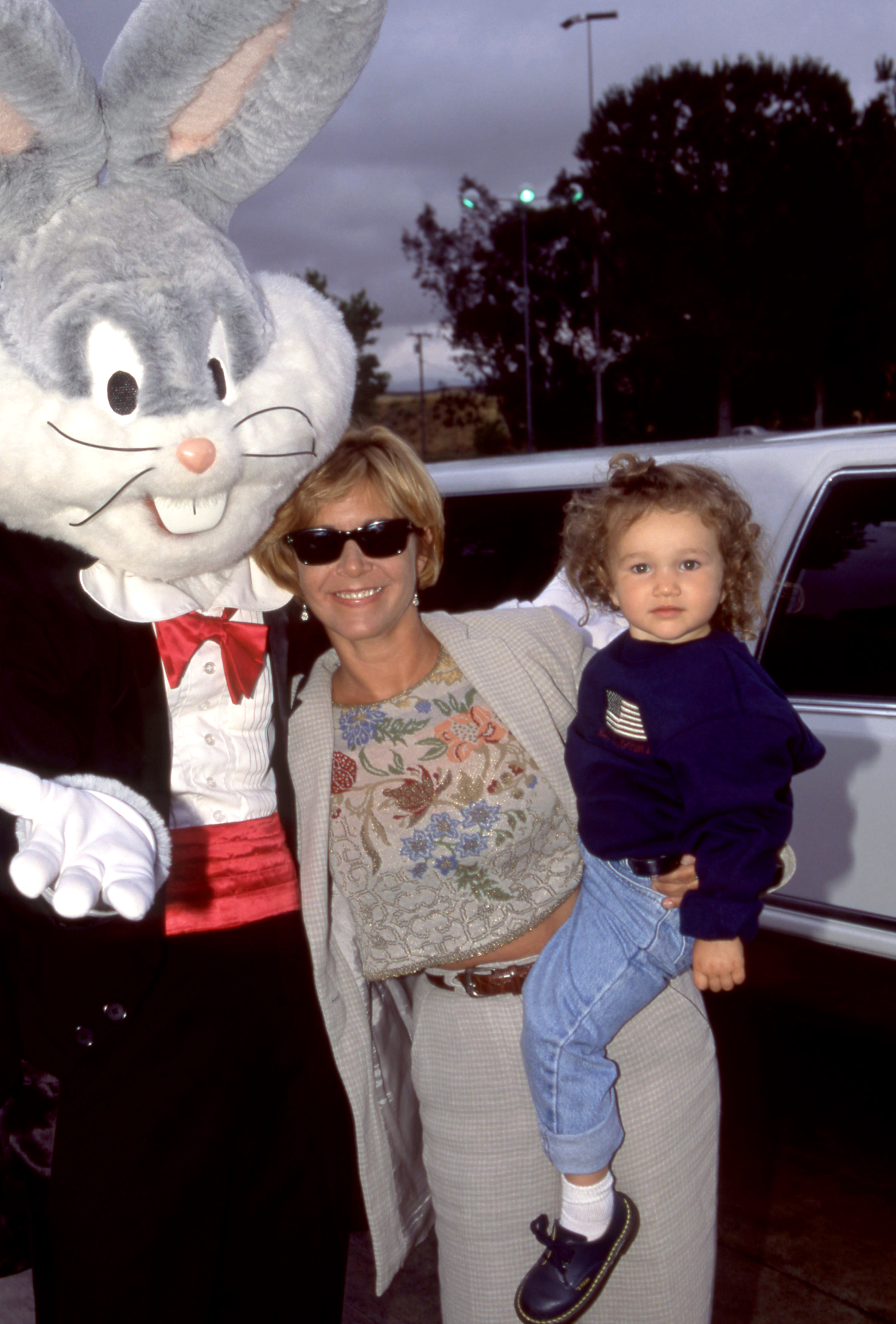 A person, wearing sunglasses and a patterned sweater, holds a child next to a person in a Bugs Bunny costume