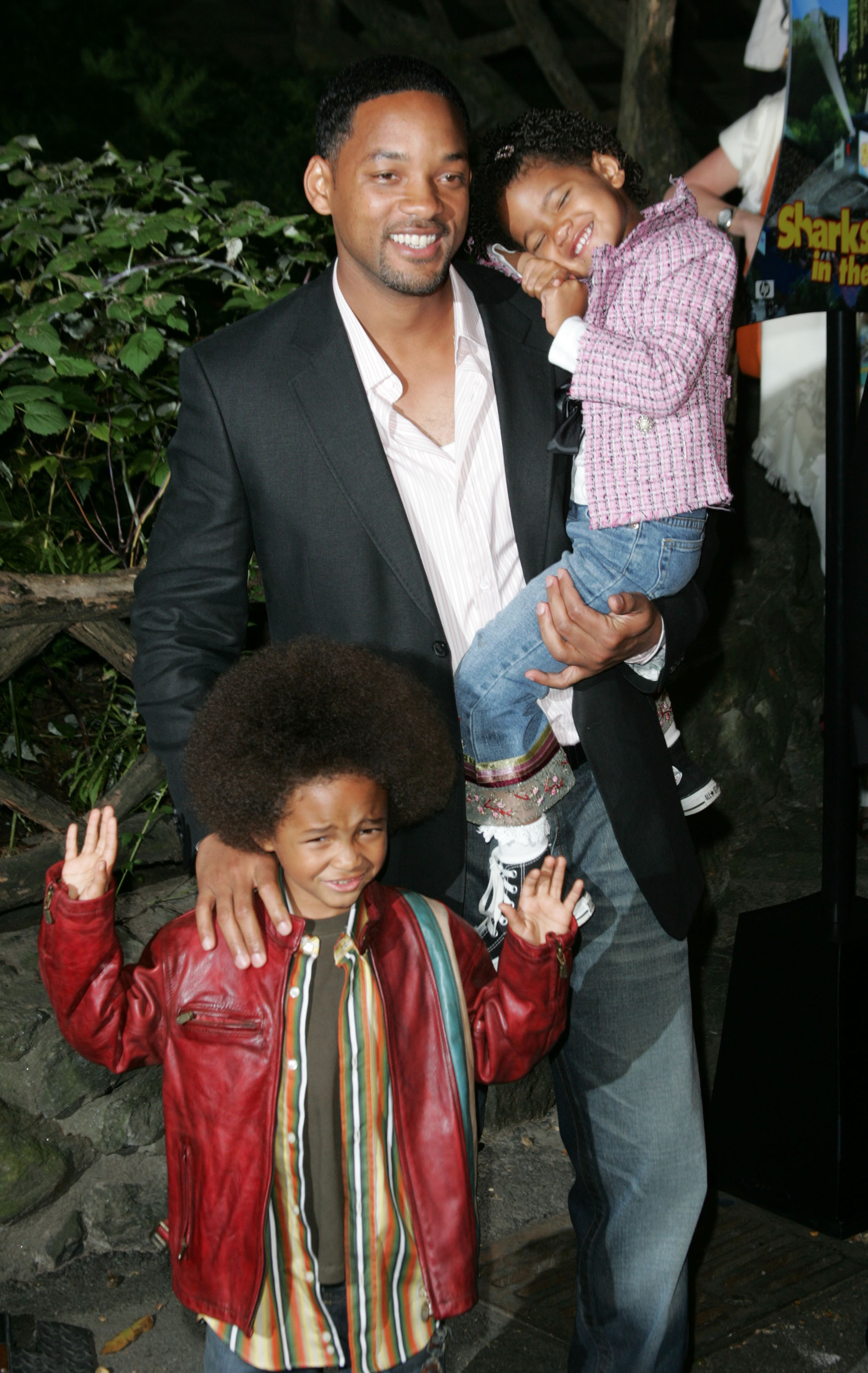 I don’t know who the people in the image are, but it shows an adult in a blazer with two joyful children. One child is wearing a red jacket, the other is being held