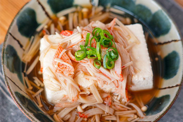 豆腐の上にエノキとカニがのった料理のクローズアップ。ネギが飾られている。