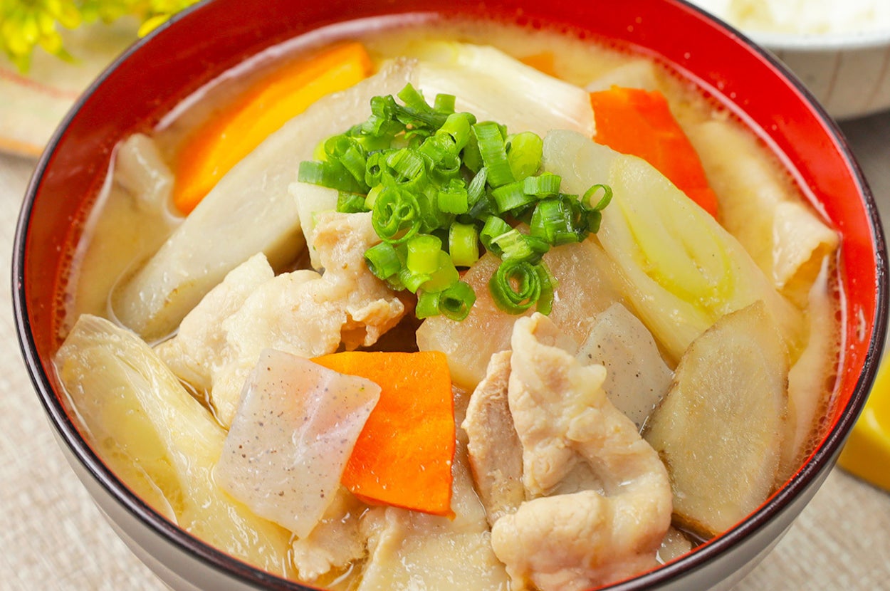 豚汁の入った器。具材には豚肉、人参、ねぎ、ゴボウ、こんにゃくが見える。背景にご飯と緑の葉がある。