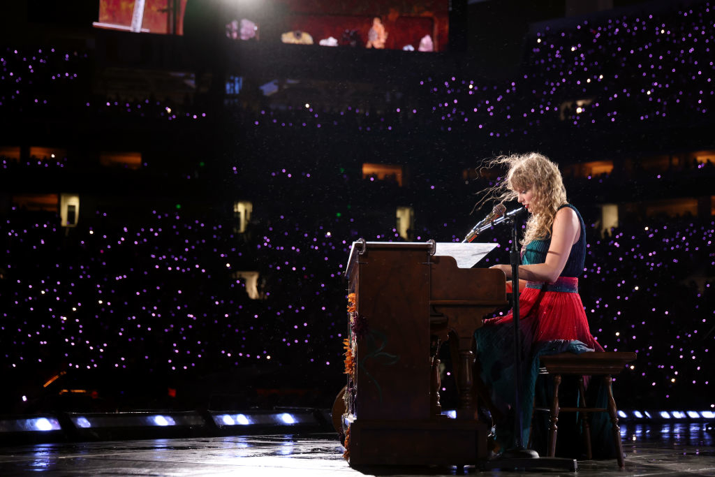 A singer performs on stage, seated at a piano, surrounded by a large, illuminated audience