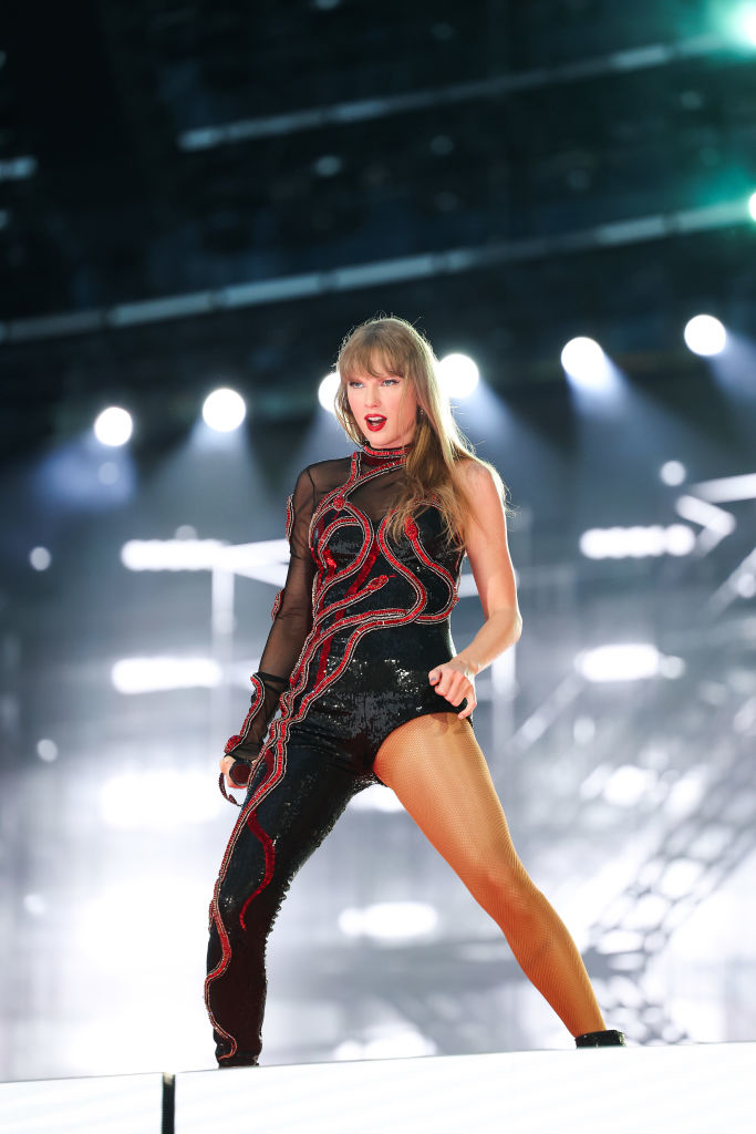 A performer, wearing a sparkling asymmetrical black and red outfit, stands on stage during a live performance