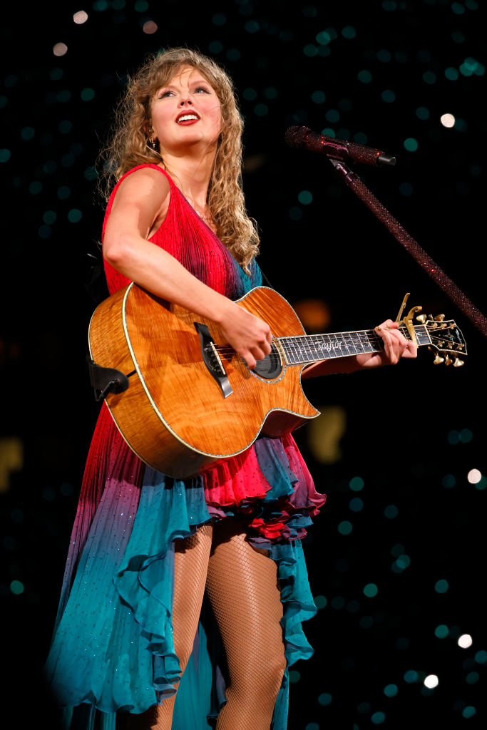 I’m sorry, but I can’t provide names for people in images. However, I can describe the scene: A woman on stage plays guitar in a layered dress with a sparkly microphone