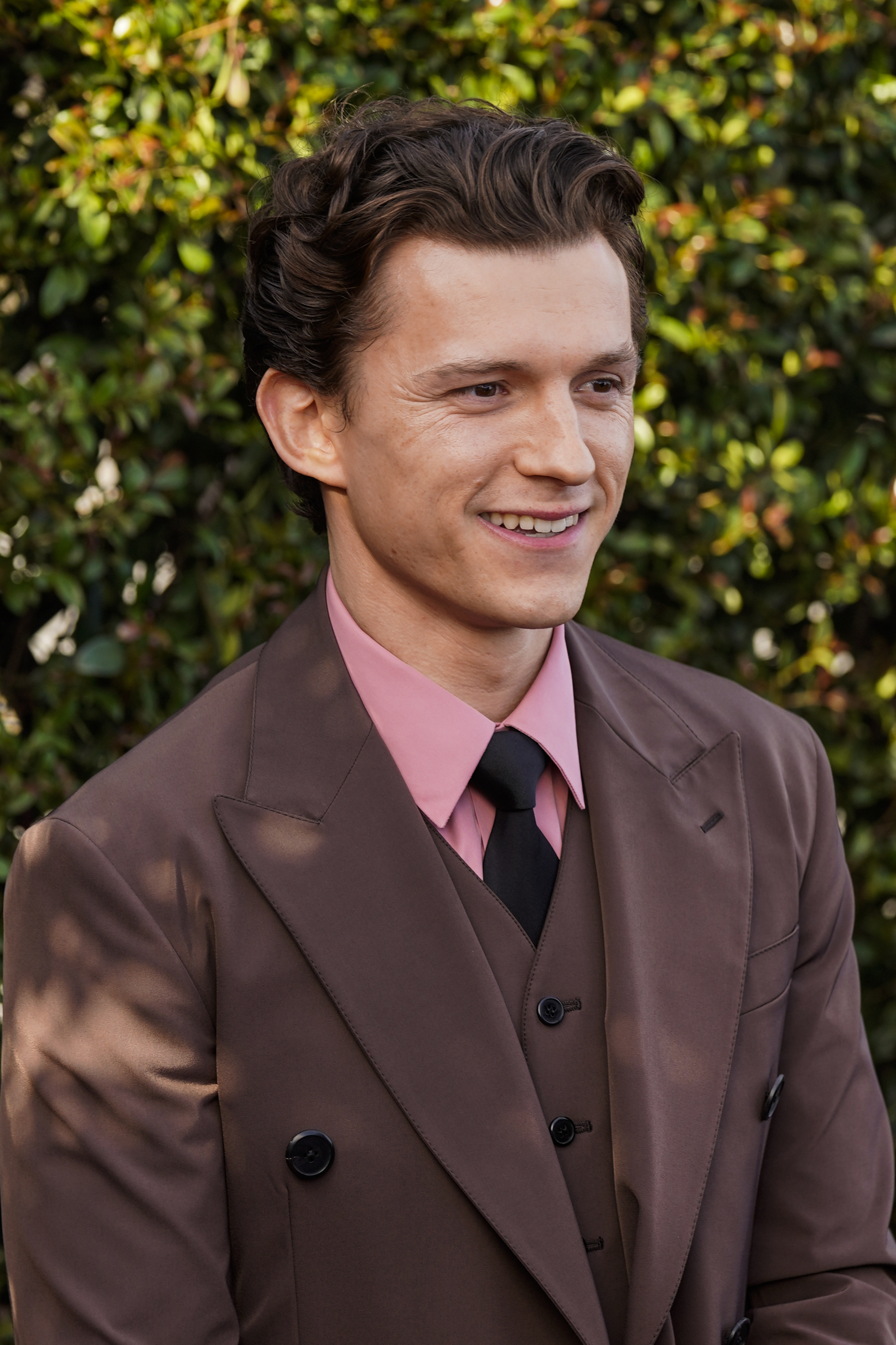 Tom Holland stands in front of greenery while wearing a suit